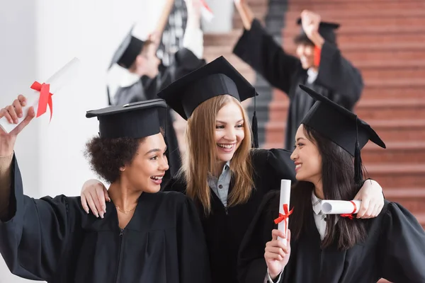 Happy interracial students with diplomas hugging — Stock Photo