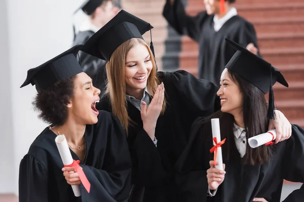 Excitados graduados multiétnicos con diplomas riendo en el salón de la universidad - foto de stock