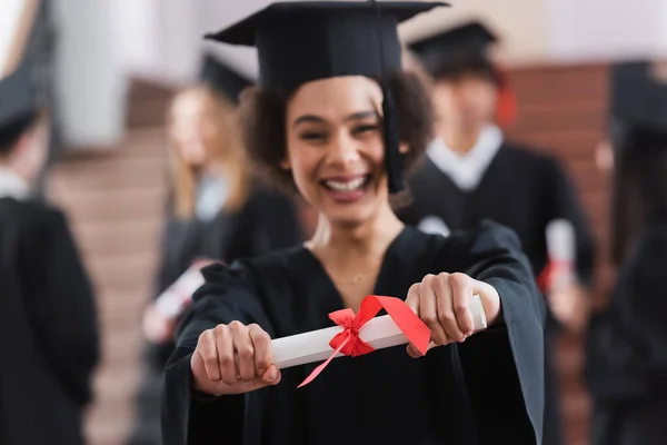 Diploma with ribbon in hands of cheerful african american student on blurred background — Stock Photo