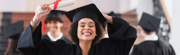 Feliz afroamericano graduado sosteniendo la tapa y el diploma, bandera - foto de stock