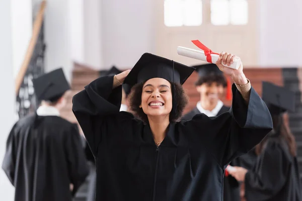 Cheerful african american graduate in academic dress holding cap and diploma — Stock Photo