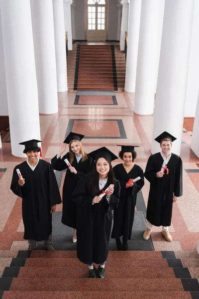 Vista ad alto angolo di scapoli multietnici sorridenti con diplomi sulle scale dell'università — Foto stock