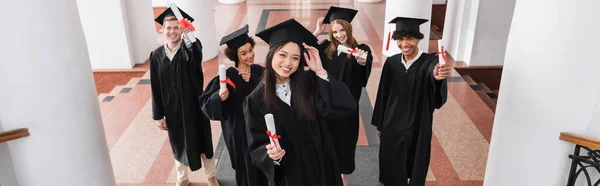 High angle view of cheerful multiethnic students holding diplomas, banner — Stock Photo