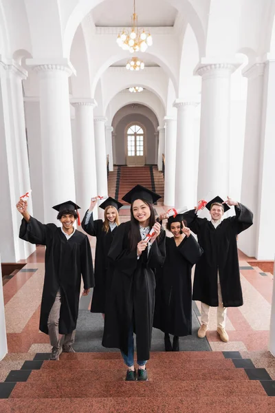 Sonriente asiático graduado celebración diploma en escaleras cerca amigos en la universidad - foto de stock