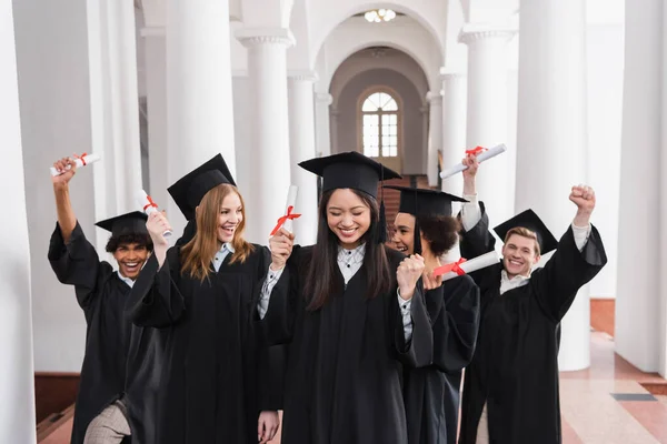 Asian graduate with diploma showing yes gesture near excited friends — Stock Photo