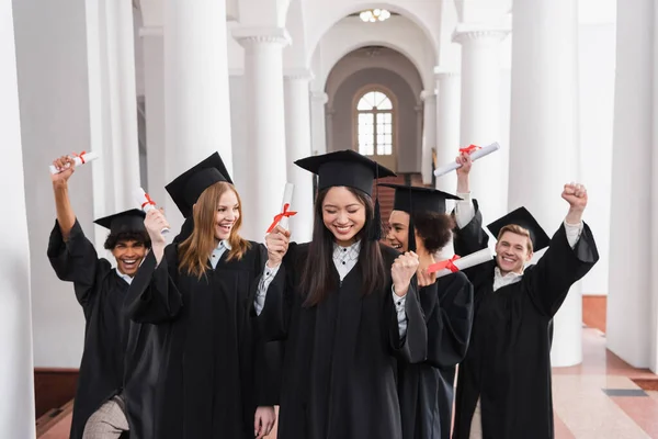 Asiático graduado con diploma mostrando sí gesto cerca emocionado amigos - foto de stock