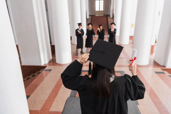 Вид ззаду випускника в академічній шапці, що тримає диплом біля друзів на розмитому фоні — стокове фото