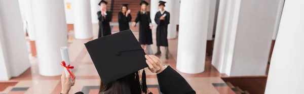 Vista posterior del estudiante en la celebración de diploma de la gorra de graduación cerca de amigos borrosos, pancarta - foto de stock