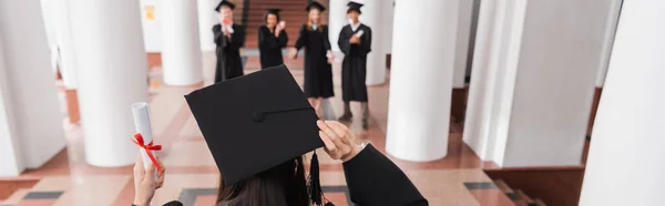 Back view of student in graduation cap holding diploma near blurred friends, banner — Stock Photo