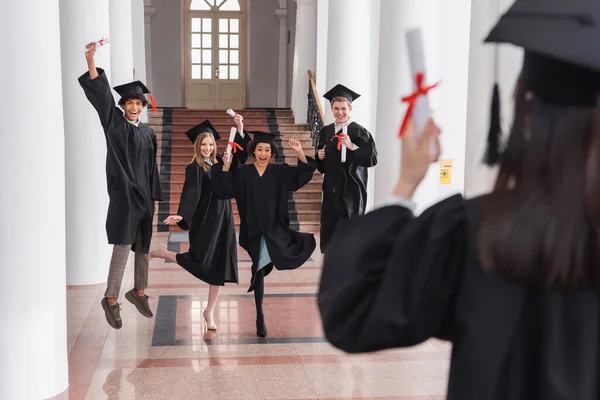 Graciosos graduados multiétnicos con diplomas cerca de un amigo en primer plano borroso - foto de stock