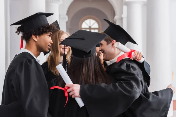 Interracial students with diplomas hugging in university — Stock Photo