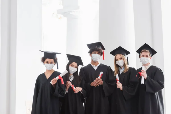 Graduados multiétnicos en máscaras médicas con diplomas - foto de stock
