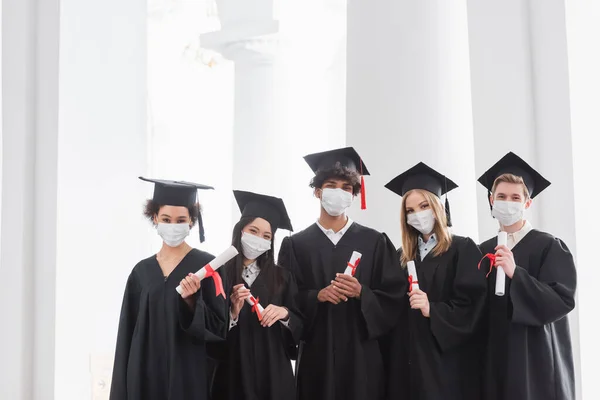 Multiethnic graduates in medical masks holding diplomas — Stock Photo