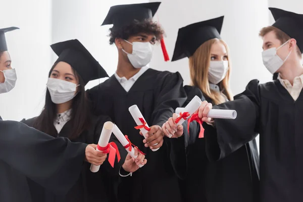 Diplomi in mani di laureati interrazziali in maschere mediche su sfondo sfocato — Foto stock