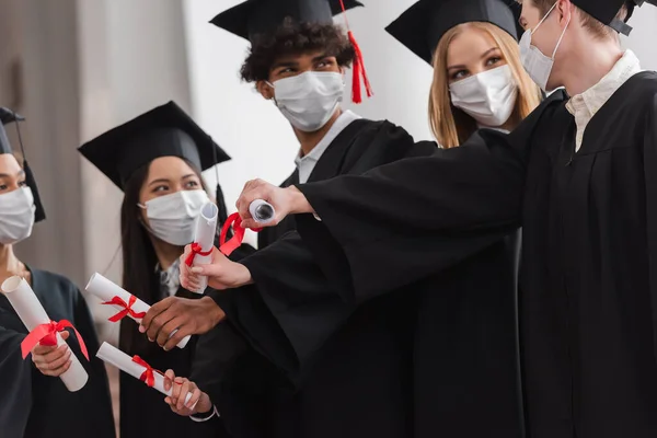 Multicultural bachelors in medical masks on blurred background holding diplomas — Stock Photo