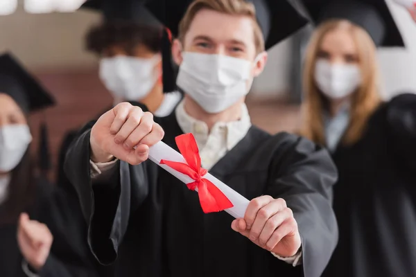 Diplôme en main du baccalauréat en masque médical sur fond flou — Photo de stock