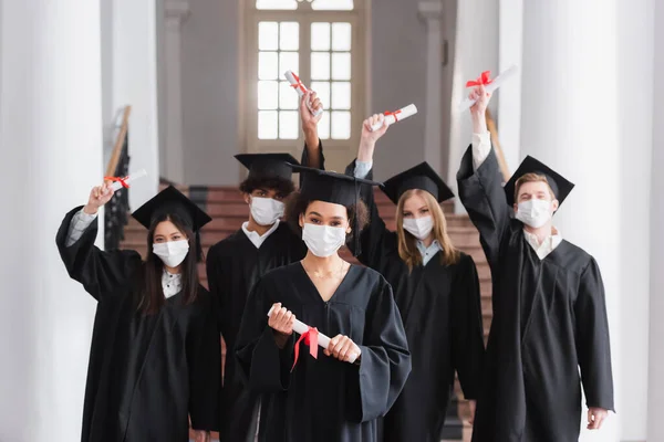 Interracial bachelors in protective masks holding diplomas — Stock Photo