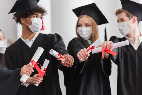 Diplomas con cintas en manos de graduados interracial en máscaras médicas - foto de stock