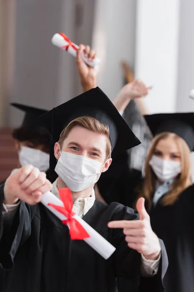 Graduado en máscara médica apuntando a diploma borroso - foto de stock