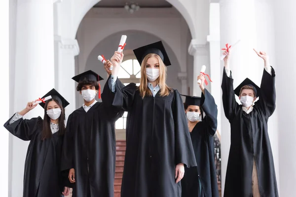 Graduados multiétnicos en máscaras médicas elevando diplomas en la universidad - foto de stock