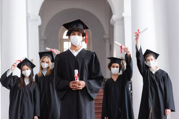 Graduado afroamericano en máscara médica con diploma cerca de amigos sobre fondo borroso - foto de stock