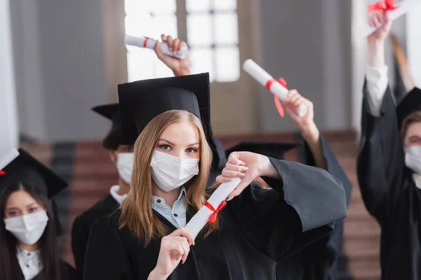 Graduado en máscara protectora con diploma cerca de amigos interracial borrosa - foto de stock