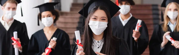 Asiática soltero en médico máscara apuntando a diploma cerca interracial amigos, bandera - foto de stock