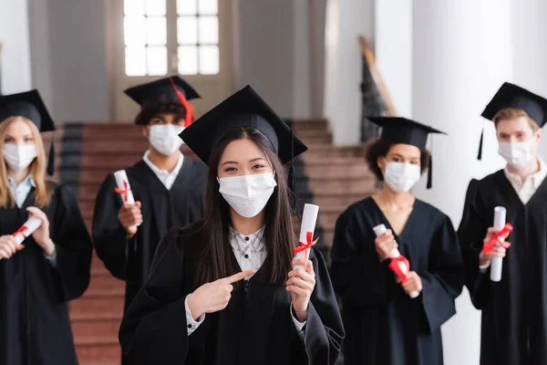 Asiática graduada en máscara médica apuntando con el dedo al diploma con cinta - foto de stock