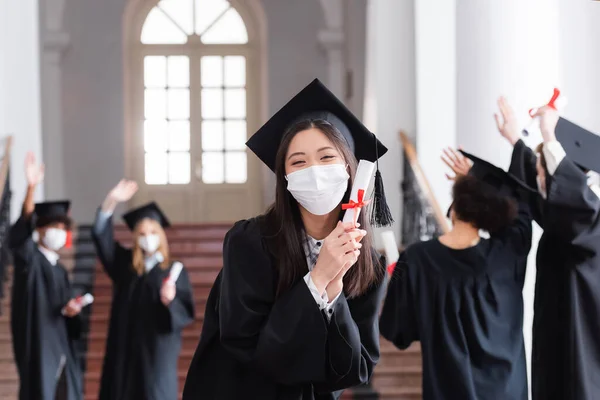 Asiática soltero en gorra y médico máscara celebración diploma cerca borrosa amigos - foto de stock