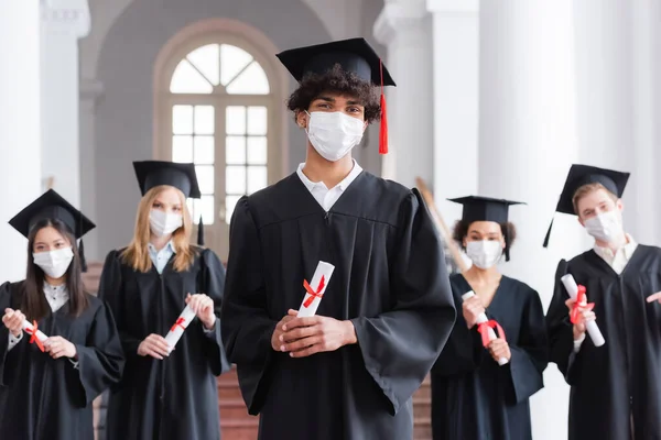 Graduado afroamericano en máscara médica y vestido académico con diploma - foto de stock