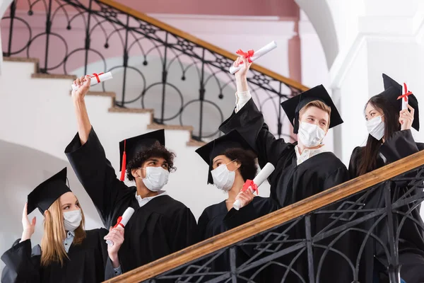 Multicultural bachelors in protective masks holding diplomas near railing in university — Stock Photo