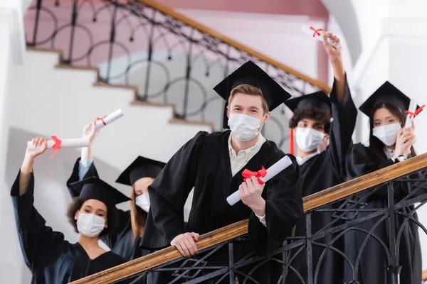 Licenciado en máscara médica con diploma cerca de amigos multiétnicos en un fondo borroso - foto de stock