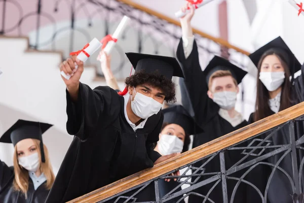 Estudiante afroamericano en máscara médica con diploma cerca de amigos sobre fondo borroso - foto de stock