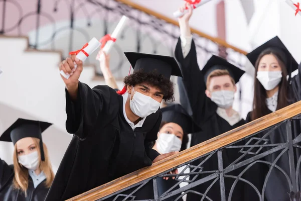 African american student in medical mask holding diploma near friends on blurred background — Stock Photo