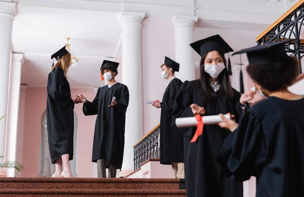 Estudantes inter-raciais em máscaras médicas e vestidos acadêmicos falando no salão da universidade — Fotografia de Stock
