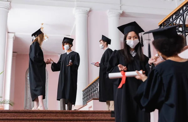 Interracial students in medical masks and academic dresses talking in hall of university — Stock Photo