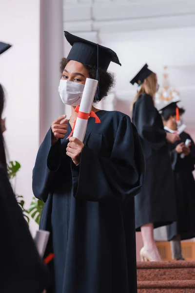 Estudiante afroamericano en máscara médica señalando diploma - foto de stock