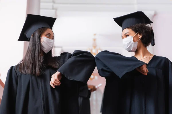 Africano americano e asiático graduados em máscaras médicas fazendo cotovelo colisão — Fotografia de Stock