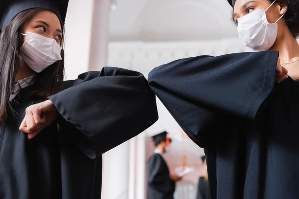 Asian and african american bachelors in medical masks doing elbow bump — Stock Photo