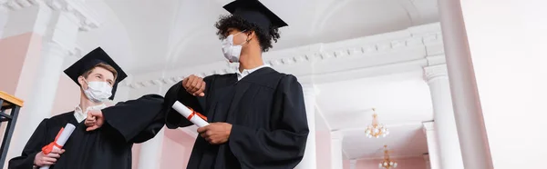 Vista de ángulo bajo de estudiantes interraciales en máscaras médicas que sostienen diplomas y hacen el golpe del codo, bandera - foto de stock