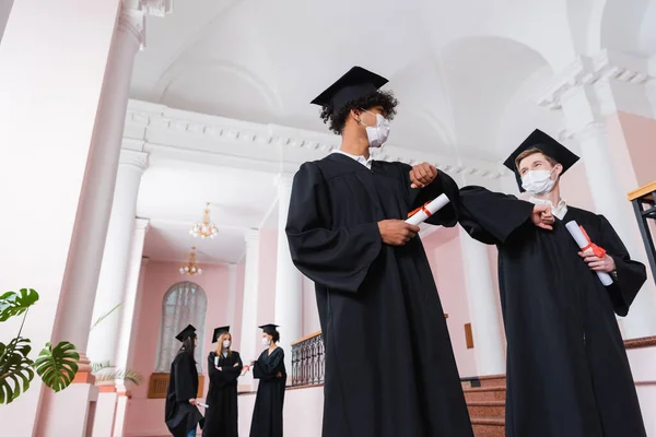 Visão de baixo ângulo de graduados multiétnicos em máscaras médicas fazendo colisão de cotovelo na universidade — Fotografia de Stock