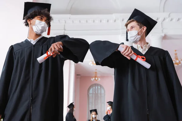 Low angle view of interracial bachelors in medical masks doing elbow bump while holding diplomas — Stock Photo