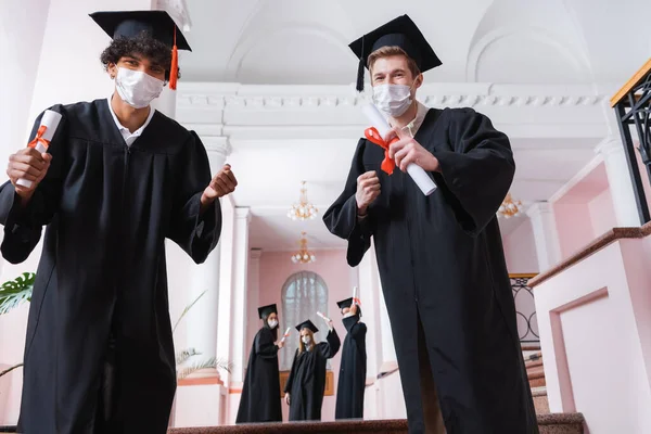 Multiethnic bachelors in medical masks showing yes gesture in hall of university — Stock Photo