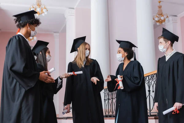 Solteros multiétnicos con máscaras médicas apuntando con las manos cerca de amigos - foto de stock