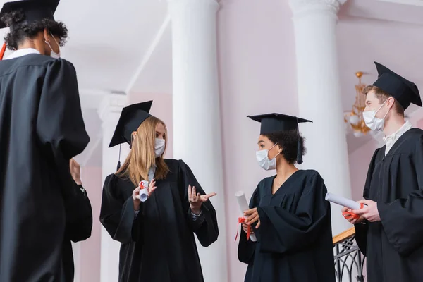 Bachelor in medizinischer Maske im Gespräch mit multiethnischen Freunden mit Diplomen in der Aula der Universität — Stockfoto