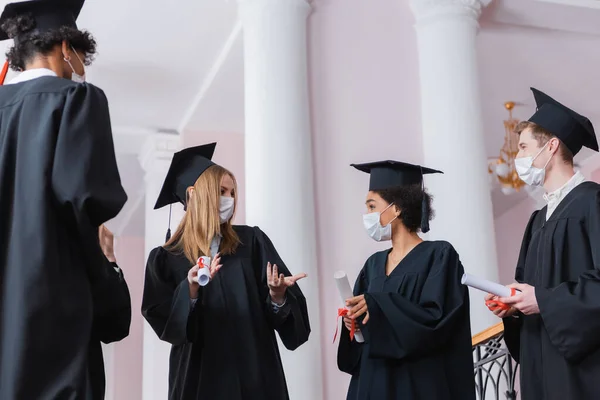 Licenciado en máscara médica hablando con amigos multiétnicos con diplomas en el salón de la universidad - foto de stock