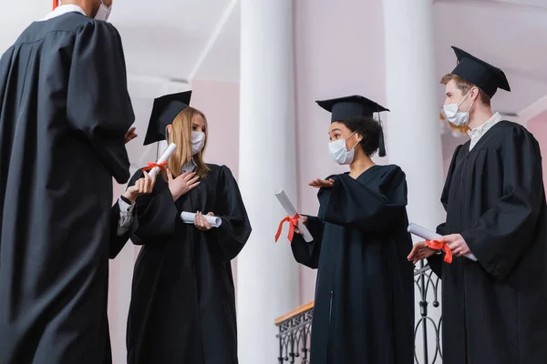 Afroamericano laureato in maschera medica che punta con mano vicino amici — Foto stock