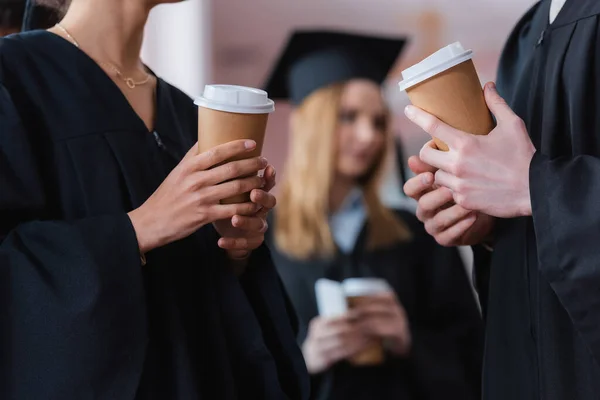 Cropped view of paper cups in hands of interracial graduates in university — Stock Photo