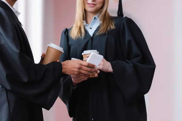 Vista recortada de graduado afroamericano sosteniendo teléfono celular cerca de amigo con café para llevar - foto de stock