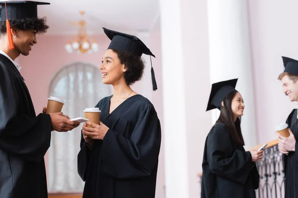 Sonriendo afroamericanos graduados con teléfonos inteligentes y papeles tazas hablando en la universidad - foto de stock